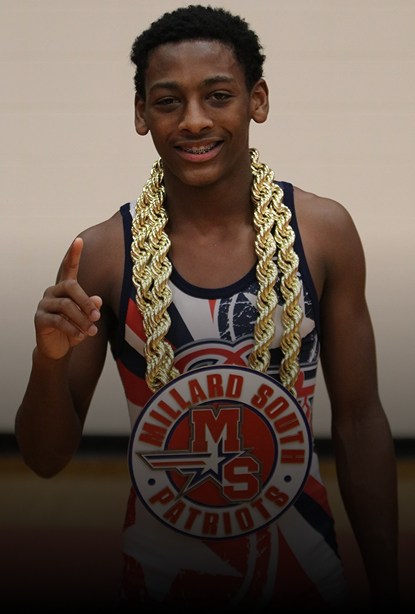 A young man wearing a two thick gold chain necklaces, standing indoors against a plain beige wall.